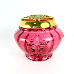 May include: A ruby-red glass jar with a decorative gold-colored metal lid. The jar has an ornate, embossed floral design and a rounded shape. The lid features an openwork pattern. The jar is likely a decorative container.