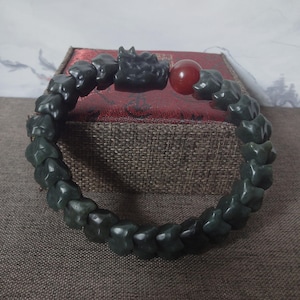May include: A dark green bracelet composed of multiple carved stone beads with a textured surface. The bracelet features a prominent carved dragon head bead and a single smooth, round, translucent red bead integrated into the circular design.