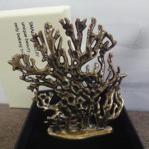 May include: A bronze-colored coral-shaped jewelry piece. The intricate design features a network of branches and holes, with a textured surface. The jewelry is displayed on a black surface, with a white box in the background that reads "SMALL JEWELRY unique beauty only belong to".