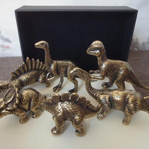 May include: A collection of five bronze-colored dinosaur figurines, including a Stegosaurus, Triceratops, and a long-necked Brachiosaurus. The detailed sculptures are displayed on a white surface, with a black box in the background.