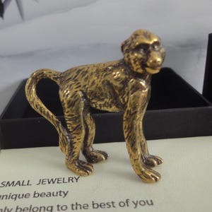 May include: A small, detailed brass monkey figurine. The monkey is standing on all fours with its tail curled. The figurine has a textured surface and is set against a black box. The text "SMALL JEWELRY unique beauty only belong to the best of you" is visible.