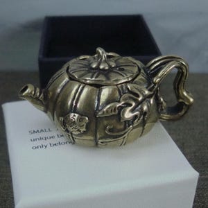 May include: A small, antique-style teapot in a pumpkin shape, crafted from a bronze-colored metal. The teapot features a lid with a stem, a curved handle, and a spout. The side of the teapot has a fish and leaf design. The text on the paper says "SMALL unique b only belong".