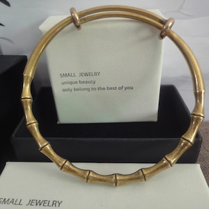 Peut inclure: Un bracelet jonc doré avec un motif bambou. Le bracelet est en métal et possède une ouverture à charnière. Le texte "SMALL JEWELRY unique beauty only belong to the best of you" est visible sur l'emballage.