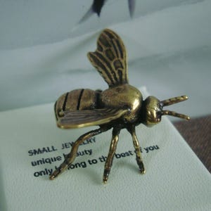 May include: A small, gold-colored bee figurine with detailed wings and legs. The bee is sitting on a white surface with the text "SMALL JEWELRY unique beauty only belong to the best of you".