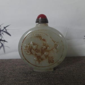 May include: A pale green, oval-shaped snuff bottle carved from stone, featuring a relief of a flowering branch and a Chinese character. The bottle is topped with a silver-toned metal cap and a smooth, rounded red stone finial at the very top.