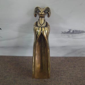 Peut inclure: Une statue de couleur bronze représentant une figure à tête de bélier portant une longue robe avec les bras croisés. La statue présente des cornes et des traits du visage détaillés. La robe descend jusqu'à la base.