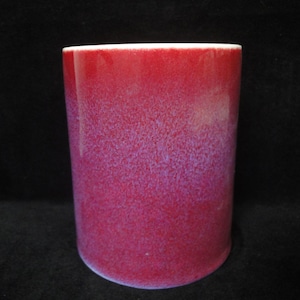 May include: A cylindrical ceramic cup with a gradient of red hues, transitioning from a deep crimson at the base to a lighter shade at the top. The cup has a white rim and a speckled texture.
