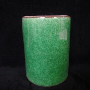 May include: A cylindrical, green ceramic cup with a crackle glaze. The cup has a smooth, rounded rim with a brown edge. The exterior of the cup is a vibrant green color with a network of fine cracks across the surface.