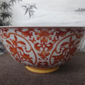 May include: A white ceramic bowl with a vibrant orange floral pattern. The bowl has a yellow base and a wide, curved shape. The intricate design features flowers and swirling vines, creating an elegant aesthetic.