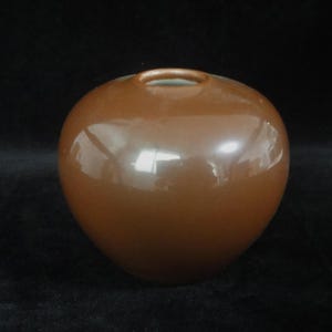 May include: A small, round, bulbous vase with a smooth, glossy, solid brown finish. The vessel features a narrow, circular opening at the top, revealing a light-colored interior rim. The surface reflects light, highlighting its curved, seamless shape.