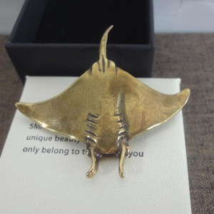 May include: A bronze-colored manta ray figurine with outstretched wings, resting on a white surface. The figurine has detailed features and is set against a black box. The text on the white surface reads "SM unique beauty, only belong to you."