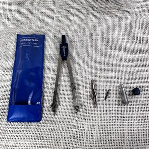 May include: A Staedtler Arco compass set featuring a blue plastic storage pouch with white text, a metal drafting compass with a blue top, an interchangeable attachment, a small lead refill, and a clear plastic container with a blue cap.