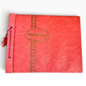 May include: A rectangular coral-colored photo album with a textured cover. A vertical gold-trimmed band features a central decorative frame containing the word Photographs in gold script. A pink cord with a tassel is threaded through the left side.