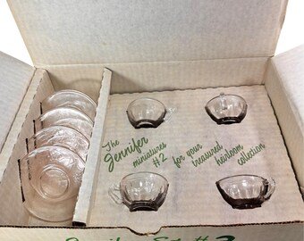 May include: A cardboard box containing a miniature tea set with four clear glass cups and four pink glass saucers. The box interior features green text reading The Jennifer miniatures #2 for your treasured heirloom collection.