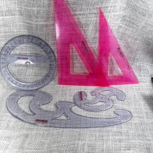 May include: A set of drafting tools including two transparent pink plastic triangles, a clear circular protractor, and a clear French curve template. Each tool features printed markings and the Koh-I-Noor brand name in red text on the surface.