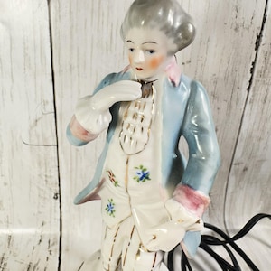 May include: A porcelain figurine of a man in 18th-century attire. The figure wears a light blue coat with pink trim, a white vest with gold accents, and a powdered wig. The figurine has a detailed face and is posed with one hand at the neck.