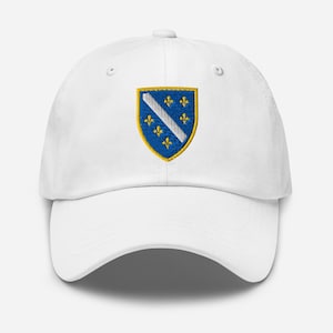 Könnte beinhalten: Eine weiße Baseballkappe mit einem gestickten Wappen auf der Vorderseite. Das Wappen ist blau mit einem gelben Rand und enthält einen diagonalen weißen Streifen sowie sechs gelbe Lilien, die zu je drei auf jeder Seite angeordnet sind.
