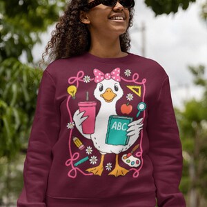May include: Burgundy sweatshirt featuring a cartoon duck wearing a pink bow and holding a drink, a book with "ABC" on it, and school supplies. The design is outlined with pink swirls and flowers. The sweatshirt is worn by a person wearing sunglasses.