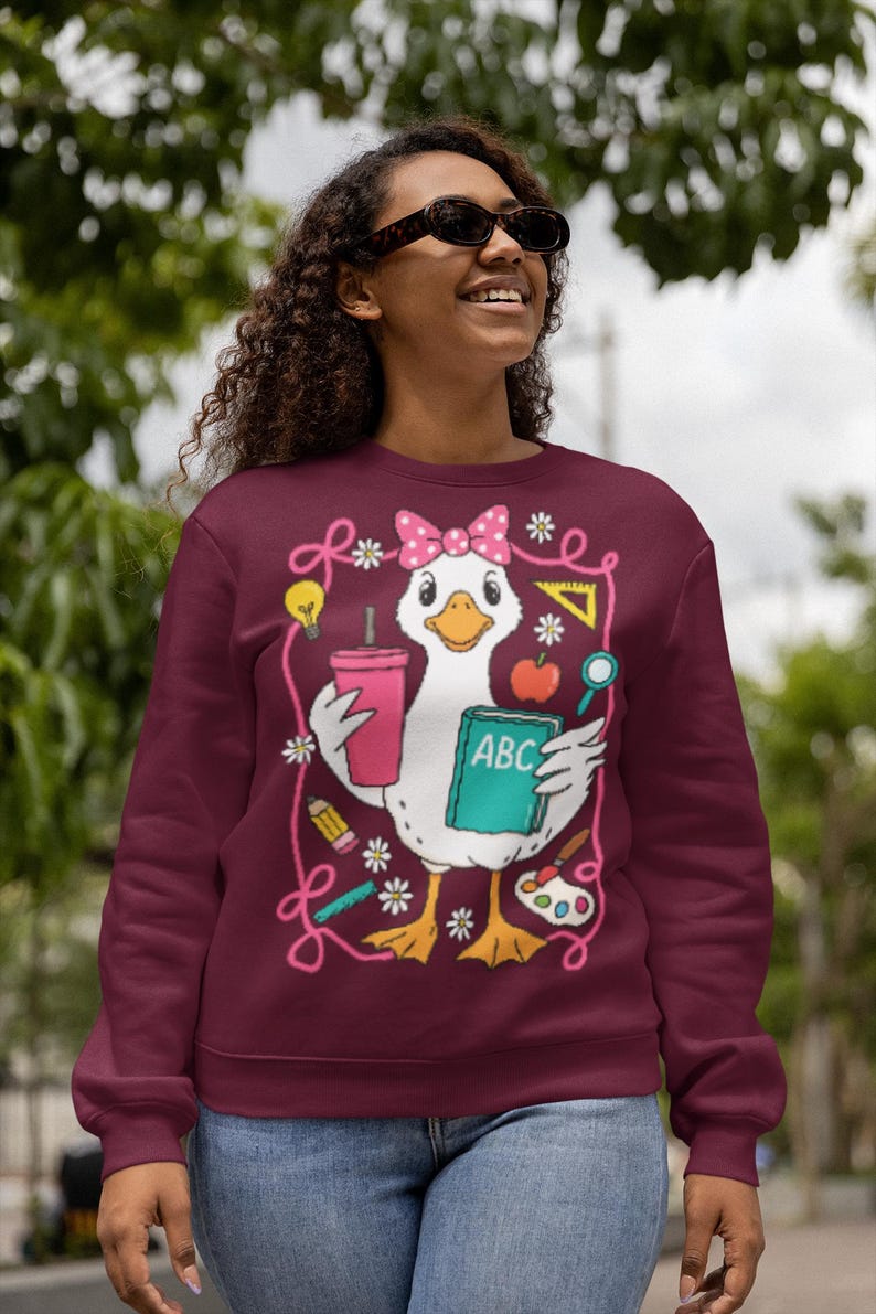 May include: Burgundy sweatshirt featuring a cartoon duck wearing a pink bow and holding a drink, a book with "ABC" on it, and school supplies. The design is outlined with pink swirls and flowers. The sweatshirt is worn by a person wearing sunglasses.