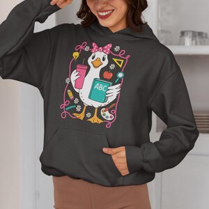 May include: Dark gray hoodie featuring a cartoon goose with a pink bow, holding a pink tumbler and a book labeled "ABC." The design includes school-themed elements like a lightbulb, apple, and paint palette, outlined in pink.