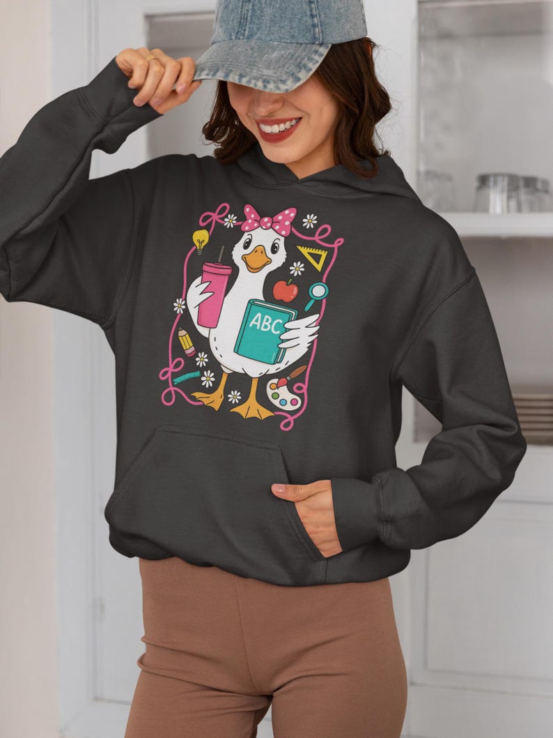 May include: Dark gray hoodie featuring a cartoon goose with a pink bow, holding a pink tumbler and a book labeled "ABC." The design includes school-themed elements like a lightbulb, apple, and paint palette, outlined in pink.