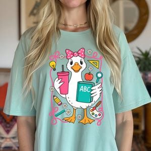 May include: A light teal t-shirt featuring a cartoon goose with a pink bow, holding a pink tumbler and a book with "ABC" on it. The design includes school-themed elements like a pencil, ruler, and paint palette.
