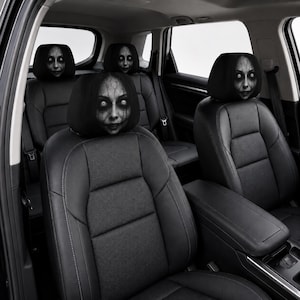 May include: A set of black car headrest covers featuring a printed graphic of a pale, wide-eyed face with dark hair and cracked skin textures. The covers are installed on the headrests of black leather vehicle seats in a car interior.