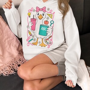 May include: White hooded sweatshirt featuring a cartoon duck wearing a pink bow and holding a pink tumbler and an ABC book. The duck is surrounded by school-themed illustrations, including a pencil, apple, and paint palette.
