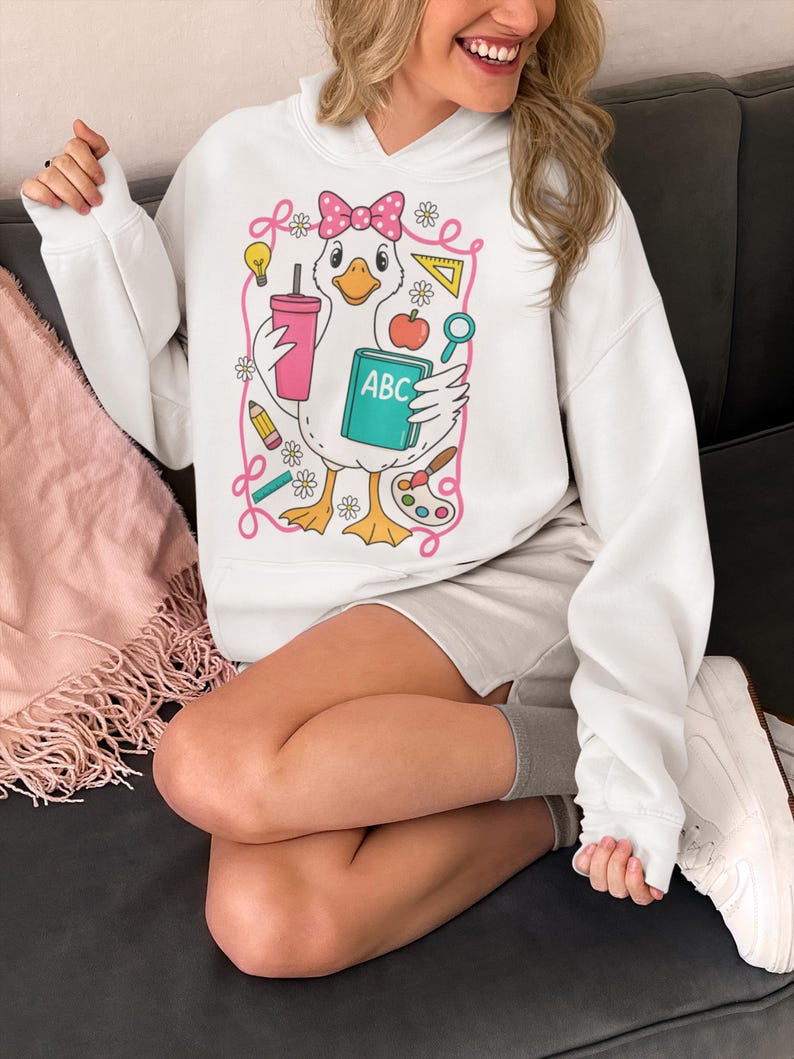 May include: White hooded sweatshirt featuring a cartoon duck wearing a pink bow and holding a pink tumbler and an ABC book. The duck is surrounded by school-themed illustrations, including a pencil, apple, and paint palette.