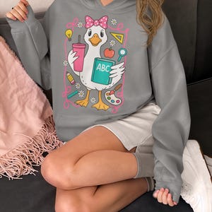 May include: Gray hooded sweatshirt featuring a cartoon goose with a pink bow, holding a drink, a book labeled "ABC", and school supplies. The design includes a pink outline with flowers and other school-themed elements. The sweatshirt is a comfortable, casual style.