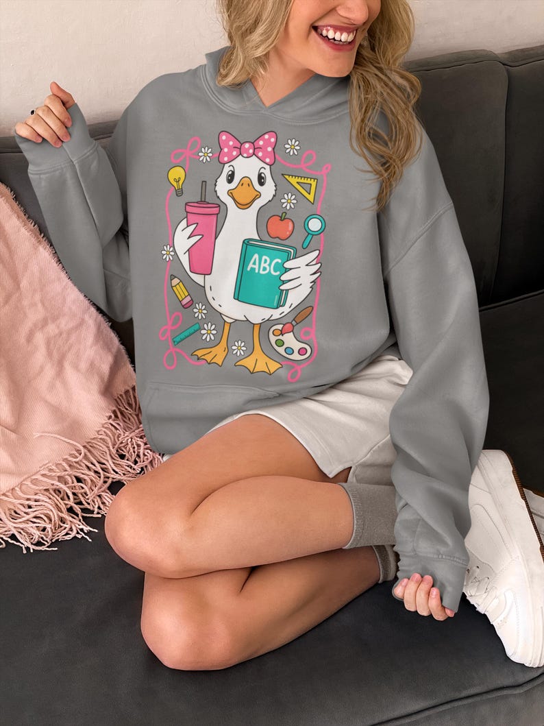 May include: Gray hooded sweatshirt featuring a cartoon goose with a pink bow, holding a drink, a book labeled "ABC", and school supplies. The design includes a pink outline with flowers and other school-themed elements. The sweatshirt is a comfortable, casual style.
