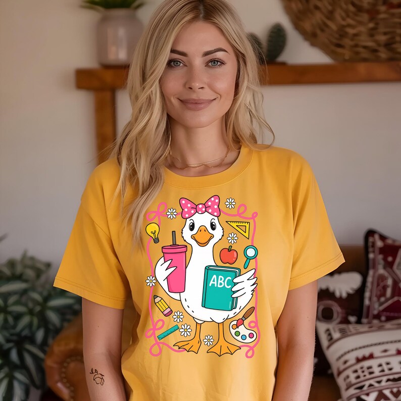 May include: Mustard yellow t-shirt featuring a cartoon goose wearing a pink bow and holding a pink cup and a book with "ABC" on it. The goose is surrounded by school-themed illustrations. The shirt is a comfortable, casual style.