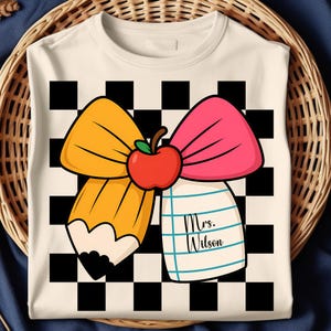 Può includere: T-shirt color crema con un design a tema scolastico. Una grande matita gialla e un fiocco rosa sono incrociati, con una mela rossa al centro. È presente anche un blocco note con il testo "Mrs. Wilson", su uno sfondo a scacchi bianco e nero.