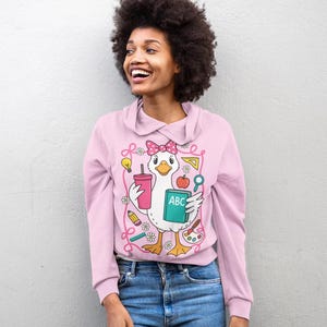 May include: A light pink sweatshirt with a cartoon duck graphic holding school supplies. The duck wears a pink bow and holds a drink, an apple, and a book with the letters "ABC". The sweatshirt has a collared neckline and long sleeves.