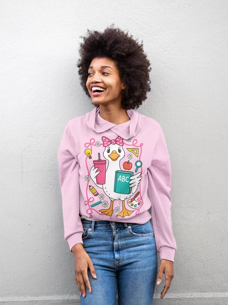 May include: A light pink sweatshirt with a cartoon duck graphic holding school supplies. The duck wears a pink bow and holds a drink, an apple, and a book with the letters "ABC". The sweatshirt has a collared neckline and long sleeves.