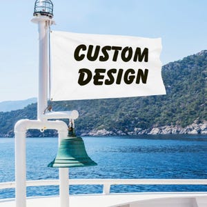 May include: A white rectangular flag featuring the black text CUSTOM DESIGN, attached to a white metal pole with a green bell mounted below the flag, positioned on a boat deck overlooking a body of water and a distant mountainous coastline.