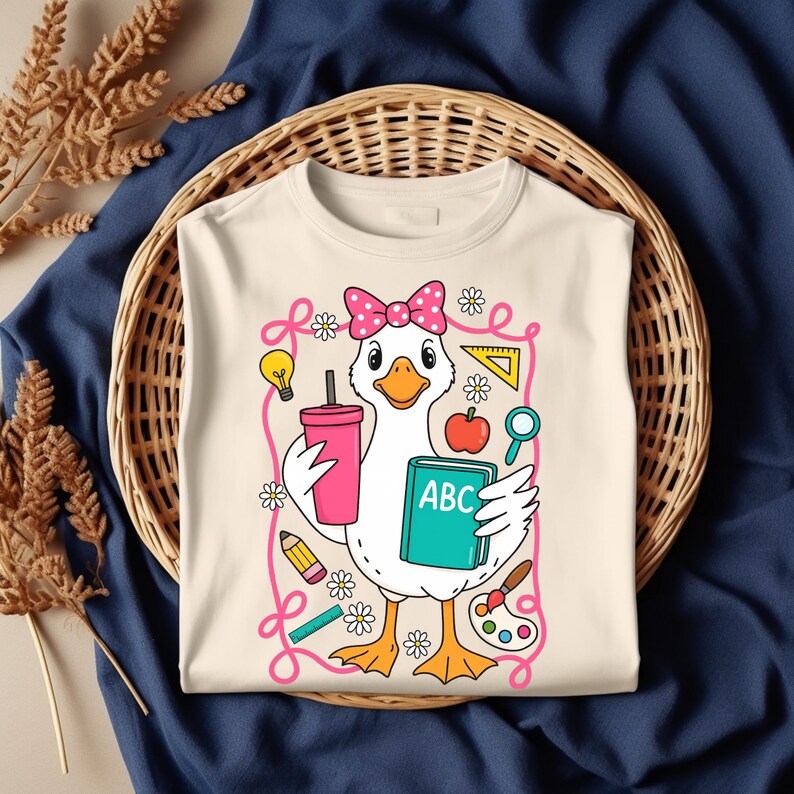 May include: Cream-colored t-shirt featuring a cartoon goose wearing a pink bow and holding a pink drink and a book labeled "ABC". The goose is surrounded by school-themed illustrations, including a pencil, ruler, and paint palette.
