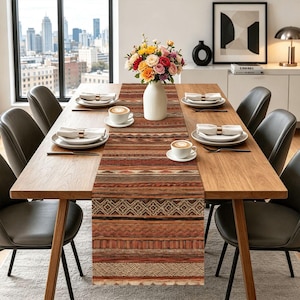 May include: A long rectangular table runner featuring a multi-colored geometric pattern in shades of rust, brown, and cream. The runner is centered on a wooden dining table set with white plates, napkins, coffee cups, and a vase of flowers.