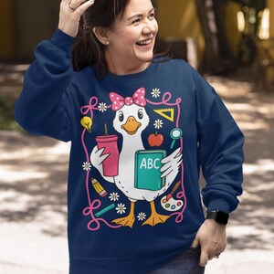 May include: Navy blue sweatshirt featuring a cartoon duck wearing a pink bow and holding a pink cup and a book with "ABC" on it. The duck is surrounded by school-themed illustrations like pencils, flowers, and a lightbulb. The sweatshirt is a comfortable, casual style.