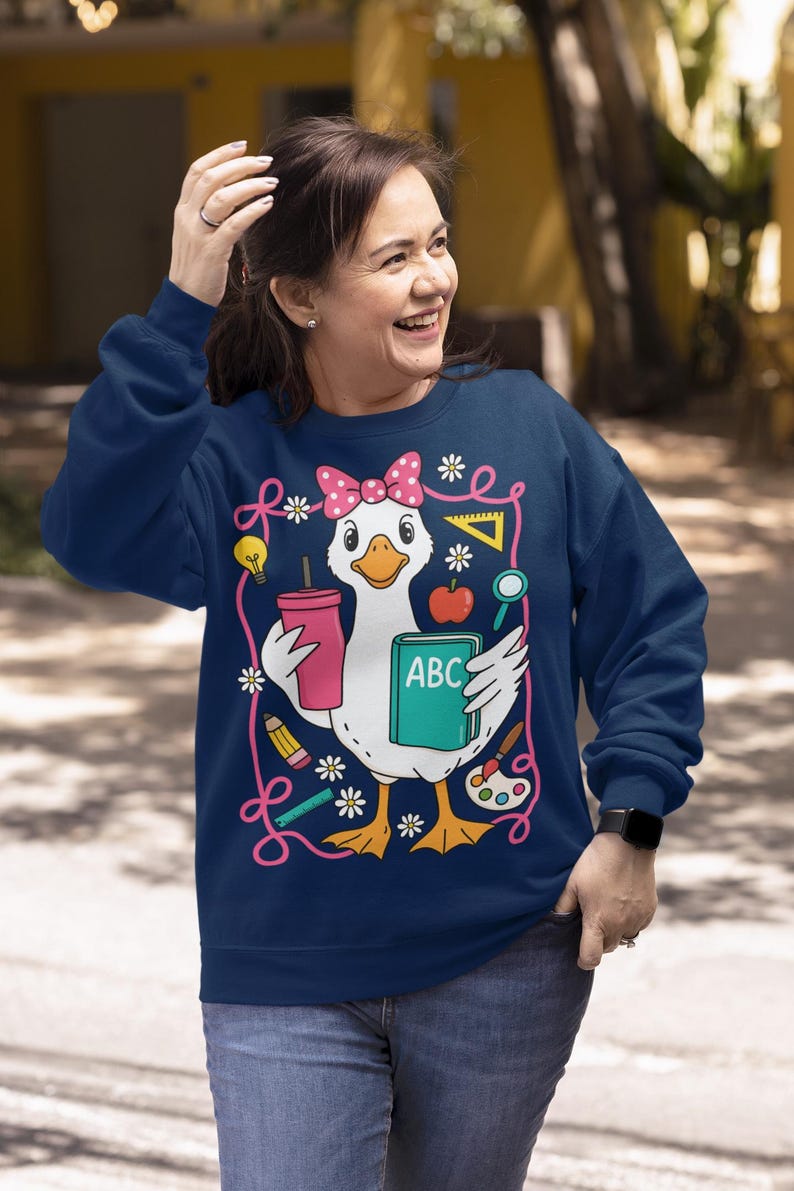 May include: Navy blue sweatshirt featuring a cartoon duck wearing a pink bow and holding a pink cup and a book with "ABC" on it. The duck is surrounded by school-themed illustrations like pencils, flowers, and a lightbulb. The sweatshirt is a comfortable, casual style.
