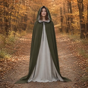 Puede incluir: Una capa con capucha que llega hasta el suelo en color verde oliva oscuro, con un forro beige claro y un cierre sencillo de lazo en el cuello. La prenda se lleva sobre un vestido a juego de color beige claro y mangas largas.