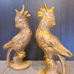 May include: Two ceramic parrot figurines. The birds are beige with gold accents on the crest, wings, and tail. They are perched on a branch-like base. The figurines are approximately 10 inches tall.