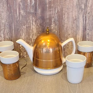May include: A copper tea set with a white teapot and four matching mugs. The teapot has a white handle and spout, and the mugs have brass handles. The set is designed for serving tea.