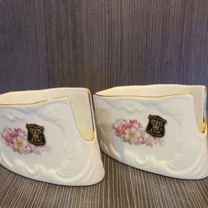 May include: Two white porcelain napkin holders with gold trim and floral designs. Each holder features a crest with a crown and the text "E&R". The holders have a curved shape and are placed on a wooden surface.