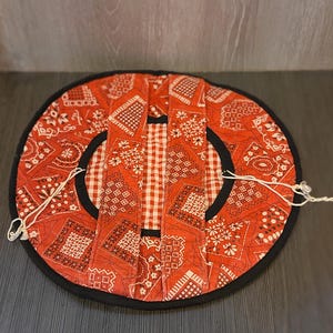 May include: A circular quilted fabric cover featuring a red and white bandana print with a central panel of red and white gingham check. The item has a black fabric border, two parallel fabric straps across the center, and white drawstring cords.