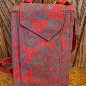 May include: A red fabric crossbody bag with a flap closure. The bag features a teal floral pattern and a matching strap. The bag is rectangular and appears to be made of a woven material.