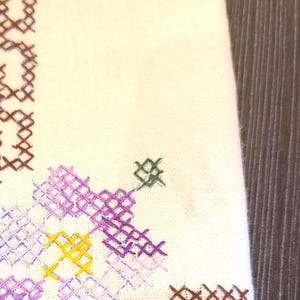 May include: Close-up of white fabric with cross-stitch embroidery. The embroidery features a floral design in shades of purple, yellow, and green, with brown accents. The background is a wooden surface.