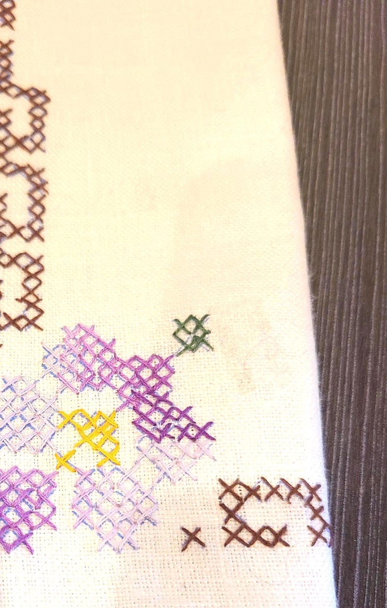 May include: Close-up of white fabric with cross-stitch embroidery. The embroidery features a floral design in shades of purple, yellow, and green, with brown accents. The background is a wooden surface.