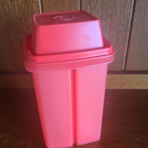 May include: A vintage, bright red plastic container with a square lid. The container has a rectangular shape with vertical grooves on the sides. The lid has a slightly raised center with a circular design. The container is likely for food storage.