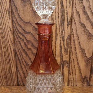 May include: A vintage glass perfume bottle with a clear, faceted stopper. The bottle has a diamond-patterned base and a reddish-brown neck. The stopper is a clear, multi-faceted crystal. The bottle is displayed on a wooden surface.